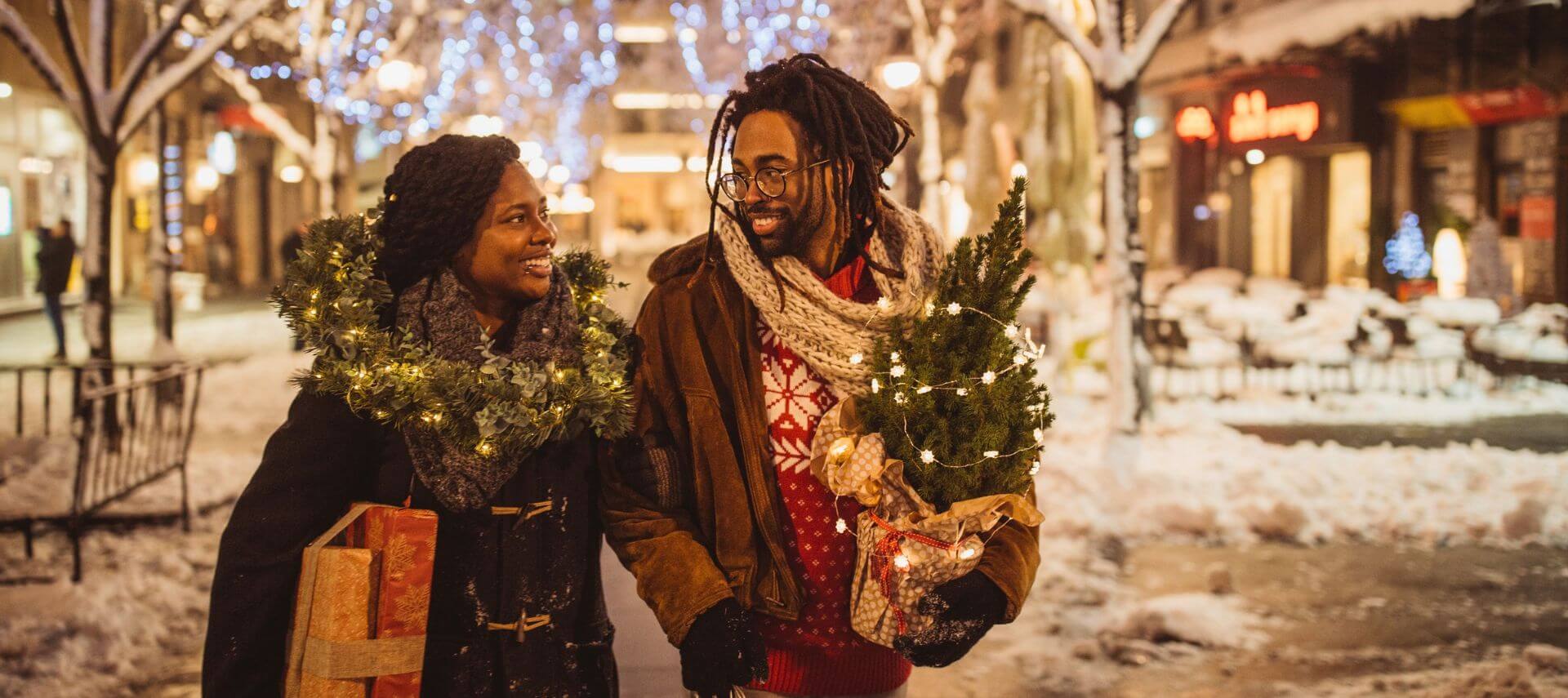 A couple smiling at each other while walking a snowy street at night, carrying a wrapped tree and gifts, with holiday lights strung overhead.