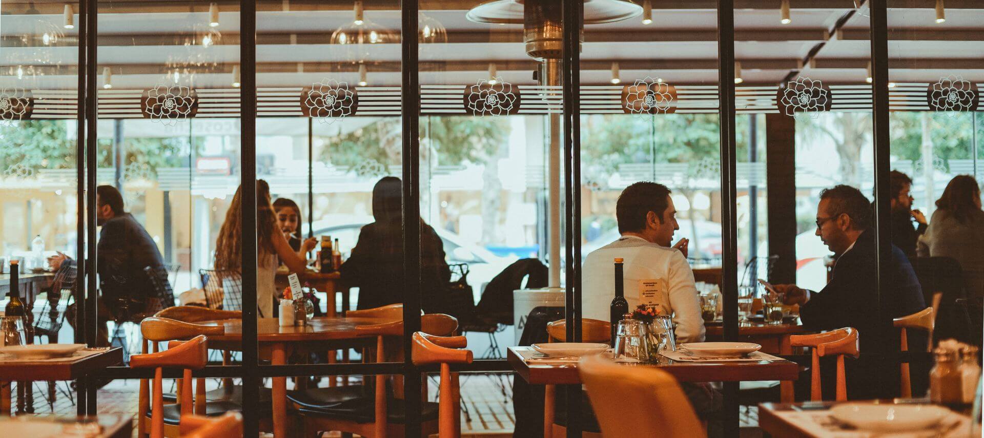 A warmly lit restaurant dining room shows guests enjoying meals and conversation through large glass windows.