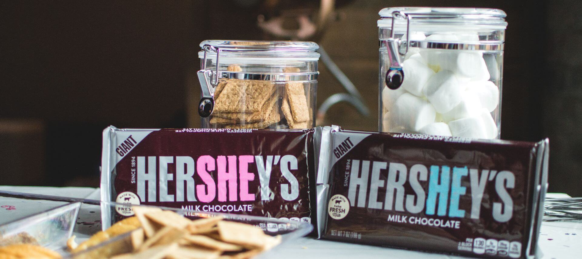 Two Hershey's milk chocolate bars sit in front of glass jars filled with graham crackers and marshmallows.