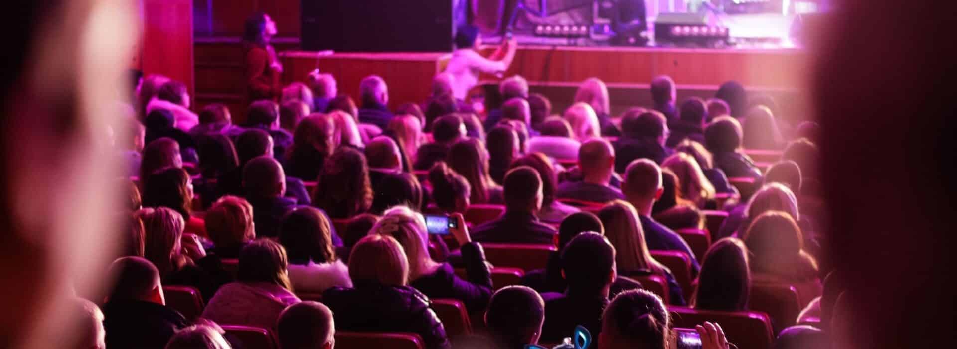 the backs of a crowd of people watching a theatre production the lighting has a pinkish purple hue to it
