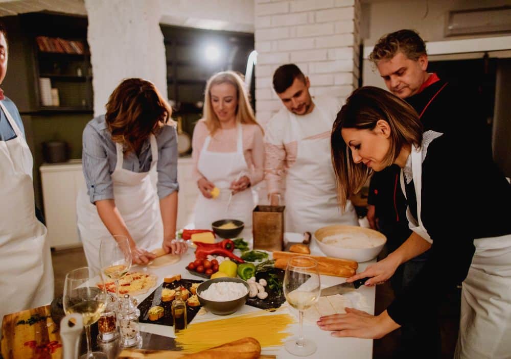 Group of people wearing white aprons in professional kitchen. Table full of veggies, pasta, bread 