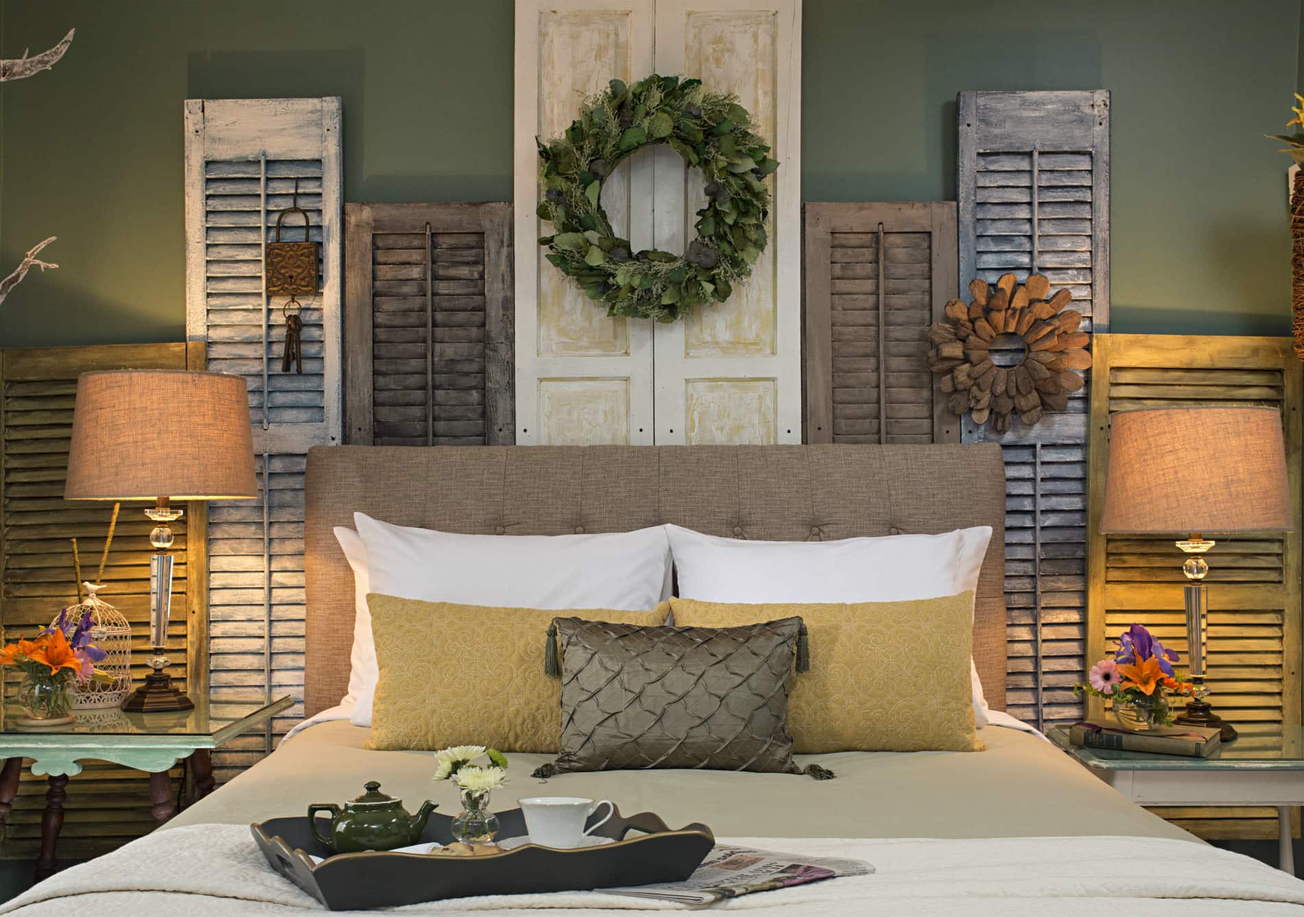 green room with bed covered in white and tan linens with golden yellow throw pillows and a decorative display of grey, brown and white window shutters behind the bed