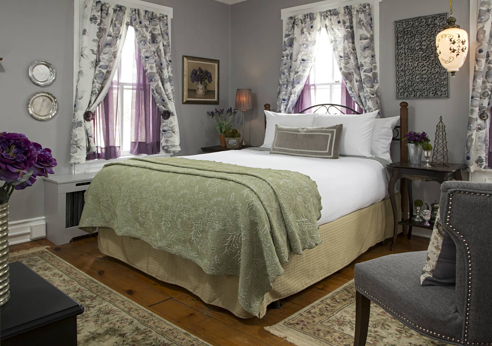 Grey room with bed covered in white linens with green quilt. Two windows by the bed have floral print curtains with purple sheers