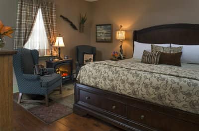 Taupe room with large bed on dark wooden frame, cream and brown floral print sheet, two grey chairs, faux-fireplace with lamp